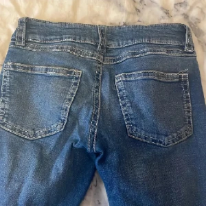 Blå jeans från Gina Tricot, storlek 170 - Snygga blå jeans från Gina Tricot med klassisk femficksdesign och dubbla silverknappar i midjan. Jeansen har tydliga kontrastsömmar och bälteshällor, perfekt för en trendig och avslappnad look.