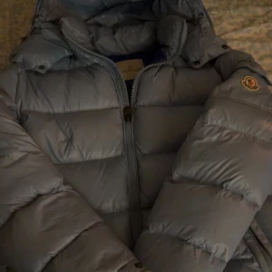 Ljusblå pufferjacka från Moncler - Säljer en snygg ljusblå pufferjacka från Moncler med huva och dragkedja. Jackan har stora, vadderade partier och klassisk Moncler-logga på ärmen. Perfekt för kalla dagar när du vill hålla stilen och värmen.