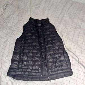 Svart dunväst från Everest - Svart quiltad dunväst från Everest med hög krage och dragkedja framtill. Storlek 146/152 Västen är lätt och smidig, perfekt att slänga över en hoodie eller tröja. Klassisk och stilren design utan synliga loggor på framsidan.