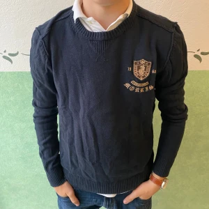 Mörkblå stickad tröja från Morris - Snygg mörkblå stickad tröja från Morris med broderad logga och emblem på bröstet. Tröjan har rund halsringning, ribbade muddar och är långärmad. Perfekt för dig som gillar klassisk och stilren preppy-look. Står L men sitter som M