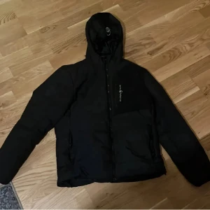 Sail Racing jacka  - Svart pufferjacka från Sail Racing med huva och dragkedja framtill. Jackan har en stilren look med logga på bröstet och praktiska fickor. Perfekt för kalla dagar och har en skön, vadderad känsla. Passar dig som gillar sportig och clean stil.