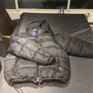 Svart pufferjacka från Polo Ralph Lauren - Svart pufferjacka från Polo Ralph Lauren i storlek XL men passar som M. Jackan har huva, två fickor med dragkedja och klassisk röd broderad logga på bröstet. Tillverkad i polyester och perfekt för kalla dagar. Snygg och stilren design med quiltade partier. Priset kan diskuteras. Först till kvarn