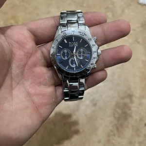 Hugo Boss klocka i stål med blå urtavla - Snygg klocka från Hugo Boss med blå urtavla och tre mindre visartavlor. Boetten och länken är i blankt rostfritt stål. Klockan har tydliga siffror och detaljerade knappar på sidan. Perfekt accessoar för dig som gillar stilrena detaljer.