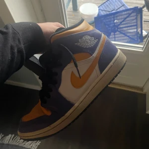 Nike Air Jordan 1 mid Lakers  - Säljer ett par Nike Air Jordan 1 mids i Lakers-färger. Skorna har lila och gula detaljer med vit bas, svart snörning och klassisk Air Jordan-logga på sidan. Ovandelen är i skinn och sulan är svart. Perfekta för dig som vill sticka ut med en ikonisk sneaker.