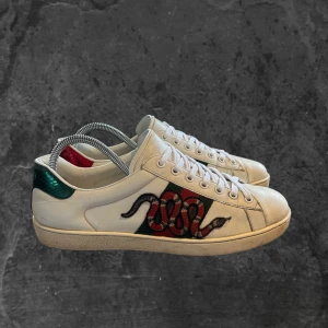 Gucci ace sneakers med ormmotiv - Säljer ett par vita Gucci sneakers i skinn med broderad röd och svart orm på sidan, gröna och röda detaljer på hälen och klassisk snörning. Snygg platt sula med unikt mönster undertill. Perfekt för dig som vill sticka ut med lyxig streetstyle.