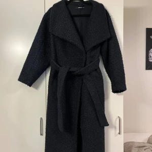 Marinblå lång kappa från Gina Tricot - Lång marinblå kappa från Gina Tricot med bälte i midjan och bred krage. Jackan har en mysig, lite fluffig struktur och är perfekt för kyliga dagar. Klassisk och stilren modell som ger en snygg siluett.