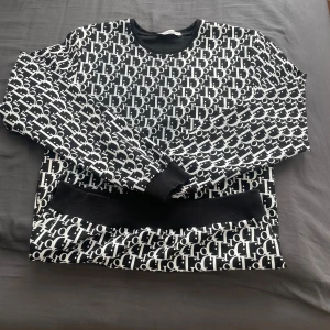 Svartvit mönstrad tröja Chiquelle - Cool svartvit tröja från Chiquelle med all-over logomönster. Tröjan har rund hals, långa ärmar och breda muddar vid ärmslut och nederkant. Perfekt statement piece för dig som gillar att sticka ut.