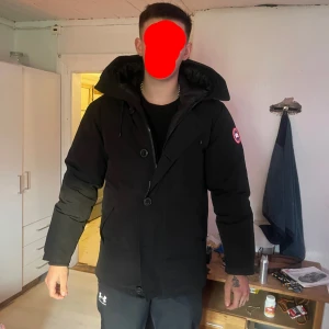 Svart dunjacka från Canada Goose - Säljer en svart dunjacka från Canada Goose med stor huva och klassisk logga på ärmen. Jackan har både dragkedja och knappar framtill, samt en snygg detalj med snörning på huvan. Perfekt för kalla dagar och riktigt stilren look.