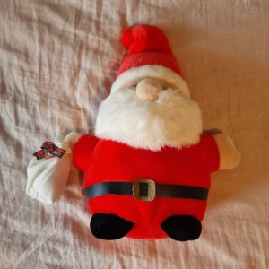 Gosedjurstomte med en säck - Klassisk rödklädd tomte i mjuk plysch, med bälte och julklappssäck. Bältet sitter inte kvar mitt på magen utan kanar ner mot fötterna. Ca 25 cm hög.