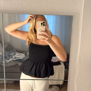 Svart volang linnetopp - Säljer en svart linnetopp med volang nedtill och smala axelband. Toppen har en croppad passform och är perfekt för dig som gillar en enkel men ändå söt stil. Materialet känns lätt och bekvämt mot huden.