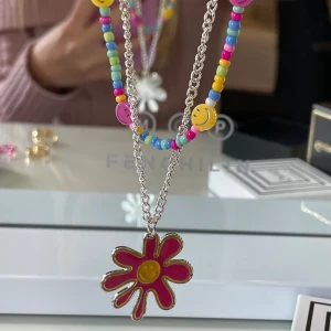 Färgglatt halsband med blomma och smileys - Dubbelhalsband med en silverfärgad kedja och en rad av färgglada pärlor och smiley-ansikten. Stort hänge i form av en rosa blomma med gul mitt. Perfekt för dig som gillar lekfulla accessoarer och färg. Säljer matchande ringar till också😊