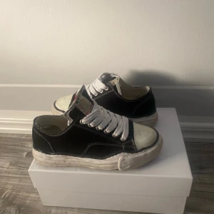  Maison Mihara Yasuhiro skor - Maison Mihara Yasuhiro sneakers i svart canvas med vita kontrastsömmar och grova vita snören. Skorna har en chunky, vit platt sula med MMY-logga på hälen och rund tå. Tungan har en tydlig etikett med röd detalj. Perfekt för dig som gillar unik streetwear. St 42