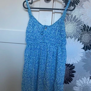 Blommig blå kortklänning - Supersöt blå och vit kortklänning från Pull&bear med blommigt mönster och smala axelband. Klänningen har en liten knytning fram och är tillverkad i mjuk bomull, perfekt för varma dagar. Sval och lätt att bära, med en avslappnad passform.
