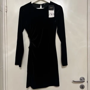 Svart sammetsklänning från Zara - Supersnygg svart sammetsklänning från Zara med lång ärm och cool detalj med glittrande paljetter i sidan och en öppning. Kort modell med rund hals och stilren siluett, perfekt för att sticka ut med en elegant touch. Festlig och trendig design som verkligen fångar blicken.