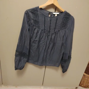 Mörkblå blus med spetsdetaljer - Säljer en mörkblå blus med långärmade ärmar och snygga spetsdetaljer på axlar och ärmslut. Blusen har rund halsringning och dekorativa sömmar framtill. Materialet känns lätt och luftigt, perfekt för dig som gillar en chill och stilren look.