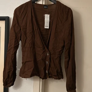 Brun omlott skjorta med knappar från Gina Tricot - Snygg brun blus från Gina Tricot med djup v-ringning och stora knappar framtill. Modellen har långärmade ärmar och markerad midja för en smickrande siluett. Perfekt att styla med jeans eller kjol för en trendig look.