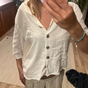 Vit skjorta från ONLY med knappar - Fräsch vit skjorta från ONLY i storlek 38, med snygga bruna knappar framtill och en avslappnad krage. Skjortan har trekvartsärm och är gjord i ett lätt, luftigt material som känns skönt mot huden. Perfekt för dig som gillar enkel och stilren look.