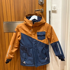 Blå och orange vindjacka med huva - Snygg tvåfärgad vindjacka från Didriksons i blått och orange. Jackan har huva, sned bröstficka, reflexdetaljer på ärmar och rygg samt dragkedja och knappar framtill. Perfekt för blåsiga dagar och har en cool, sportig look.