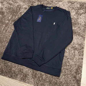 Mörkblå sweatshirt från Polo Ralph Lauren - Snygg mörkblå sweatshirt från Polo Ralph Lauren med klassisk rund hals och broderad logga på bröstet. Tröjan har ribbade muddar vid ärmslut och nederkant. Perfekt för en clean och stilren look. Storlek XL men passar även L