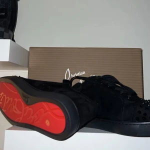 Christian Louboutin svarta sneakers med nitar - Svarta sneakers från Christian Louboutin med ikonisk röd sula och svarta nitar på tån. Skorna har snörning och är tillverkade i mocka för en lyxig känsla. Perfekt för dig som vill sticka ut med edgy detaljer och exklusiv design.
