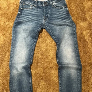 Blå jeans från Jack & Jones - Snygga blå jeans från Jack & Jones med klassisk femficksdesign och ljusa slitningar framtill. Jeansen har rak passform och normal midja, tillverkade i mjukt denimtyg för en bekväm känsla. Perfekta för dig som gillar en stilren och avslappnad look.