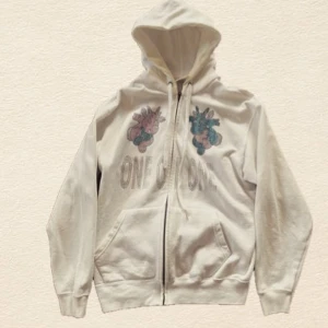 ONE OF ONE Hoodie Medium - Vit hoodie med dragkedja och huva från One Of One. Framsidan har ett tryck med två färgglada figurer och texten 'ONE OF ONE'. Tröjan har kängurufickor och ribbade muddar. Mindre avfärgning.