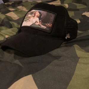 Svart Maggiore keps med mesh och tryck - Svart keps från Maggiore med mesh på sidorna och baksidan. Framtill finns ett stort tygmärke med ett retroinspirerat tryck av en kvinna i vit klänning. Kepsen har böjd skärm och justerbar baksida. Snygg metallogo på sidan.