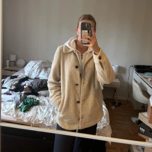 Beige teddyjacka med knappar - Mysig beige teddyjacka med klassisk krage och stora bruna knappar framtill. Jackan har två fickor på sidorna och är tillverkad i fluffigt teddy-material som ger en chill och trendig look. Perfekt för lager-på-lager och passar till de flesta outfits. Nopprigt/använt skick i tyget på vissa ställen, se sista bilden. Utöver det bra skick.