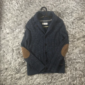 Mörkblå stickad kofta från Pier One - Snygg mörkblå stickad kofta från Pier One med ribbat mönster och bruna mockadetaljer på armbågarna. Koftan har knäppning framtill och en klassisk krage, perfekt för lager-på-lager-stil under kyliga dagar. Använd 1 gång 