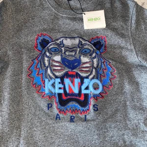 Kenzo ( äkta )  - Grå sweatshirt från Kenzo med ikonisk broderad tiger i blått, rött och svart framtill samt texten 'KENZO PARIS'. Tröjan har rund hals, ribbade muddar och lång ärm. Perfekt statement-plagg med coolt motiv och snygga färgdetaljer.