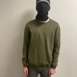 Olivgrön sweatshirt från C.P. Company - Säljer en olivgrön sweatshirt från C.P. Company med rund hals och lång ärm. Tröjan har en cool detalj med en svart lins på vänster ärm och en ficka med dragkedja. Materialet är mjuk bomull och passformen är normal. Perfekt för dig som gillar streetwear.