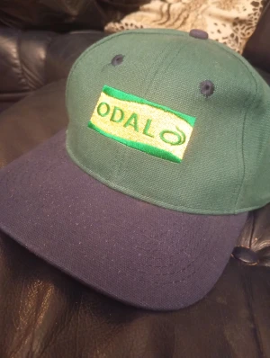 Odal keps - Snygg keps från Odal med grön krona och mörkblå skärm. Framtill finns ett broderat Odal-märke i grönt och gult. Kepsen har klassisk form med ventilationshål och är tillverkad i bomull. Perfekt för dig som vill ha en chill och stilren look.