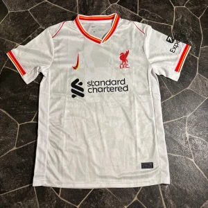 Liverpool fotbollströja Nike - Säljer en vit Liverpool fotbollströja från Nike med röd och gul kant på krage och ärmslut. Tröjan har korta ärmar, broderat klubbmärke och Nike-logga på bröstet samt sponsortryck. Materialet är lätt och andas, perfekt för match eller träning.