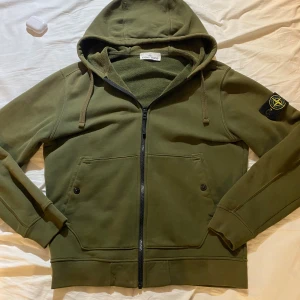 Grön hoodie från Stone Island - Säljer en grön hoodie från Stone Island med huva och dragkedja framtill. Tröjan har långa ärmar, två fickor med knappar och den klassiska Stone Island-loggan på vänster ärm. Perfekt för dig som gillar streetwear och snygga detaljer.