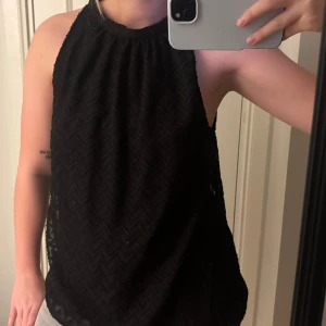 Svart ärmlös topp från Vila - Säljer en svart ärmlös topp från Vila med hög hals och snyggt diskret mönster i tyget. Toppen har en lös passform och knäpps i nacken med en liten öppning bak. Perfekt för dig som gillar stilrena och trendiga plagg.