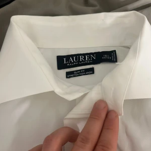 Vit skjorta från Ralph Lauren - Säljer en stilren vit skjorta från Ralph Lauren. Skjortan har en slim fit passform och är tillverkad i stretchmaterial som inte behöver strykas. Perfekt för en elegant look med klassiska knappar framtill och långärmad design. Säljer pågrund av att det är för stor storlek och kan inte använda den så har aldrig ens burit den men fick den som present