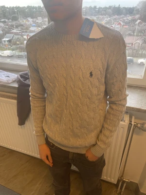 GråHelt ny stickad tröja från Polo Ralph Lauren med prislapp - Snygg grå stickad tröja från Polo Ralph Lauren med klassiskt kabelmönster och broderad logga på bröstet. Tröjan har rund hals och långa ärmar med ribbade muddar. Perfekt för dig som gillar stilrena och tidlösa plagg.