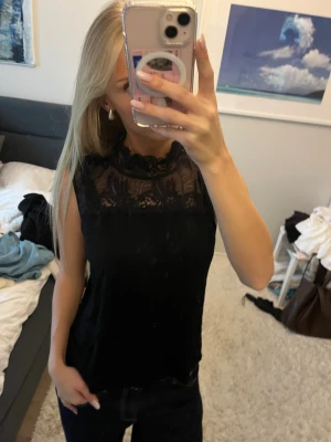 Svart blus med spetsdetaljer - Säljer en svart blus med snygga spetsdetaljer upptill och vid halsen. Blusen är ärmlös och har en hög hals med dekorativ spets som ger en cool och feminin vibe. Perfekt att styla med jeans eller kjol för en trendig look.