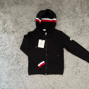 Svart Moncler hoodie med röd/vita detaljer - Svart hoodie från Moncler med huva och dragkedja framtill. Huva och ärmslut har snygga ränder i rött, vitt och grått. Moncler-logga på vänster ärm. Tröjan har fickor framtill.