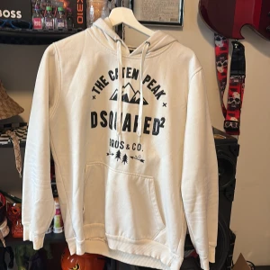 Vit hoodie från Dsquared2 med tryck - Säljer en vit hoodie från Dsquared2 i storlek L med svart tryck på bröstet och coolt bergmotiv. Tröjan har huva med snörning, stor magficka och ribbade muddar vid ärmslut och nederkant. Perfekt för dig som gillar streetwear och snygga loggor.