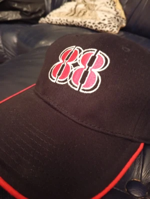 Svart keps med röd 88-brodyr - Svart keps med broderad 88 i rött och vitt framtill. Skärmen har en snygg röd kant som ger extra attityd. Klassisk form med böjd skärm, perfekt för dig som gillar sportig och cool stil. Materialet känns som bomull och kepsen har en stilren look.