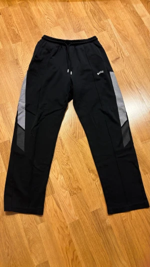 Svarta mjukisbyxor från BOSS, M - Svarta mjukisbyxor från BOSS med vita logotypen på vänster ben och coola grå mönstrade paneler längs sidorna. Byxorna har elastisk midja med snörning och är tillverkade i mjukt bomullsmaterial. Perfekt för chill eller träning.