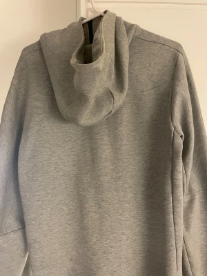 Grå Nike tech ziphoddie - Snygg grå nike tech med svart logga på bröstet och svarta detaljer. Jackan har huva, hel dragkedja framtill och en extra ficka med dragkedja på ärmen. Perfekt för dig som gillar sportig stil och vill ha något bekvämt och funktionellt. 