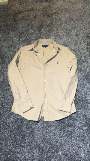 Beige skjorta Polo Ralph Lauren  - Beige långärmad skjorta från Polo Ralph Lauren i slim fit. Klassisk krage, knappar framtill och blå broderad logga på bröstet. Tillverkad i mjuk bomull, perfekt för en clean och stilren look.