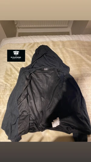 Svart vindjacka från EA7 Emporio Armani - Svart vindjacka från EA7 Emporio Armani med huva och dragkedja framtill. Jackan har meshfoder på insidan och logga på bröstet. Perfekt för dig som vill ha en stilren och sportig look. Lätt och smidig att bära.