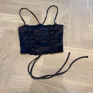 Svart korsett-topp med snörning - Snygg svart korsett-topp med smala axelband och croppad passform. Toppen har glansigt tyg och cool snörning i ryggen som ger en edgy vibe. Perfekt att styla med höga jeans eller kjol för en trendig look. Materialet känns mjukt och har en lätt satin-finish.