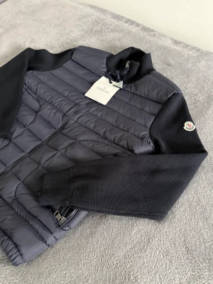 Moncler cardigan  - Snygg svart quiltad jacka från Moncler med stickade ärmar och klassisk logga på vänster ärm. Jackan har dragkedja framtill, ståkrage och två fickor med dragkedja. Perfekt för dig som vill ha en stilren och exklusiv look.