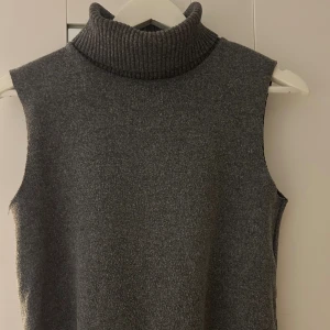 Grå ärmlös polotopp i stickad material - Säljer en stilren grå polotopp i stickad material. Toppen är ärmlös med en hög, ribbad polokrage som ger en cool och modern vibe. Perfekt att styla lager på lager eller bära som den är för en clean look.