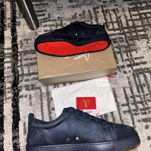 Christian Louboutin svarta sneakers mocka - Snygga svarta sneakers från Christian Louboutin i mjuk mocka med klassisk röd sula och diskret broderad logga på hälen. Skorna har rund tå, platt sula och svarta snören. Perfekta för dig som vill ha en lyxig och stilren look.