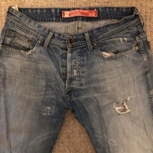 Blå straight jeans med slitningar - Säljer ett par blå jeans med straight fit och låg midja. Jeansen har slitna detaljer och hål på ena låret för en cool, avslappnad look. Klassisk femficksmodell med knappgylf och lätt tvättad finish. Perfekt för dig som gillar streetstyle och baggy jeans. Obs observera hålet på sista bilden